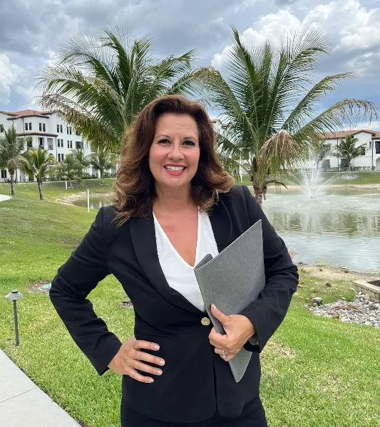 A woman in a black blazer and white top stands outdoors holding a grey folder, with palm trees and a fountain in the background.
