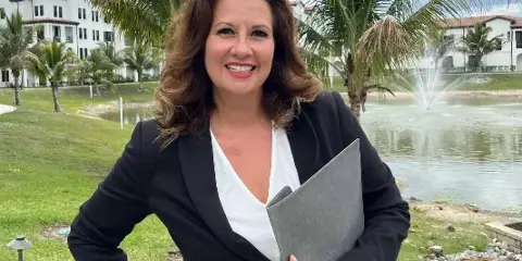 A woman in a black blazer and white top stands outdoors holding a grey folder, with palm trees and a fountain in the background.
