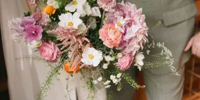 A bride and groom holding a large, colorful wedding bouquet featuring pink, white, and purple flowers.