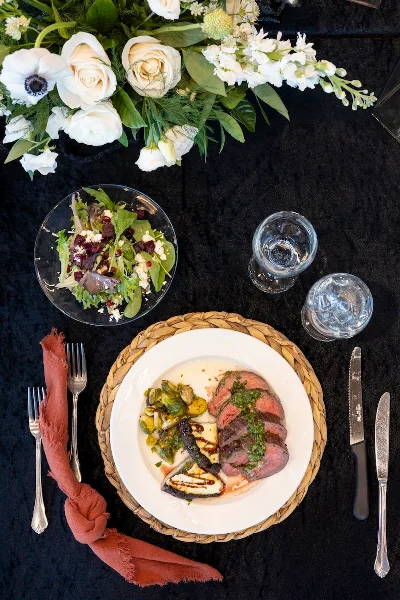Overhead view of a plated catering meal with sliced steak, brussels sprouts, and a side salad on a black tablecloth with white floral centerpiece and silverware.