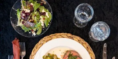 Overhead view of a plated catering meal with sliced steak, brussels sprouts, and a side salad on a black tablecloth with white floral centerpiece and silverware.