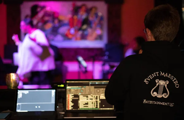 A DJ and AV technician wearing an Event Maestro Entertainment hoodie operates a mixing console and laptop at a live event.