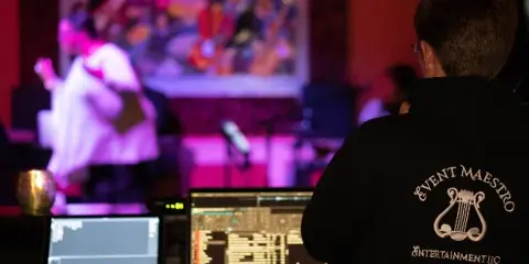 A DJ and AV technician wearing an Event Maestro Entertainment hoodie operates a mixing console and laptop at a live event.