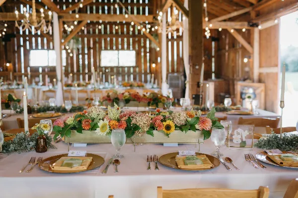 Interior of a rustic barn wedding venue with wooden beams, chandeliers, string lights, and a decorated reception table with floral centerpiece.