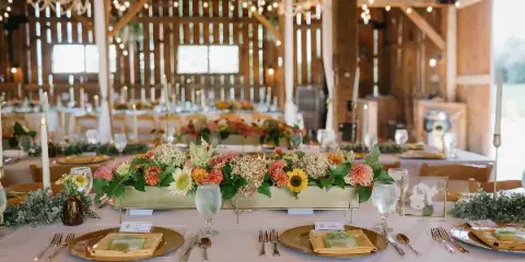 Interior of a rustic barn wedding venue with wooden beams, chandeliers, string lights, and a decorated reception table with floral centerpiece.