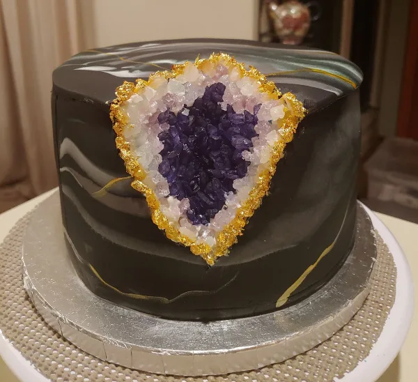 A custom black cake featuring a large, edible crystal geode design with purple and white crystals and gold accents.
