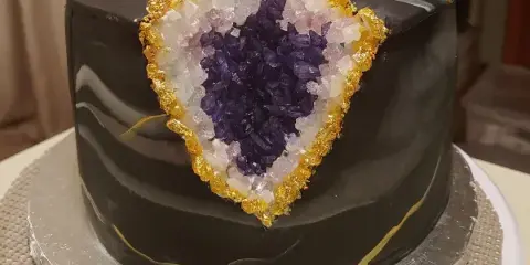 A custom black cake featuring a large, edible crystal geode design with purple and white crystals and gold accents.
