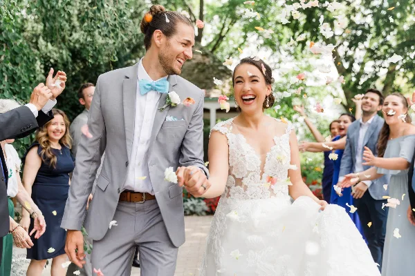 A bride and groom laughing and holding hands while walking through a crowd of guests throwing flower petals.
