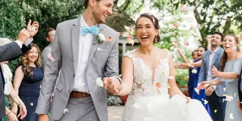 A bride and groom laughing and holding hands while walking through a crowd of guests throwing flower petals.