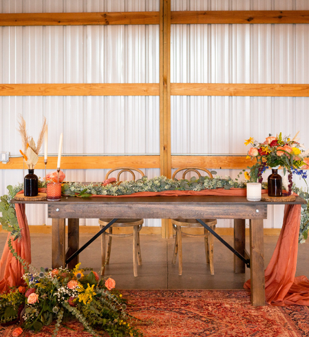 A rustic wooden table set with a long floral runner, candles, and autumn-colored floral arrangements in a barn-like venue.