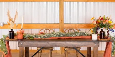A rustic wooden table set with a long floral runner, candles, and autumn-colored floral arrangements in a barn-like venue.
