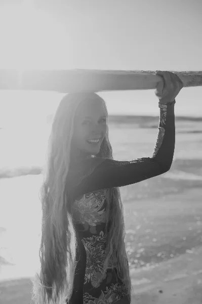 Black and white portrait of a smiling woman with long hair standing on a beach, holding onto a wooden railing.