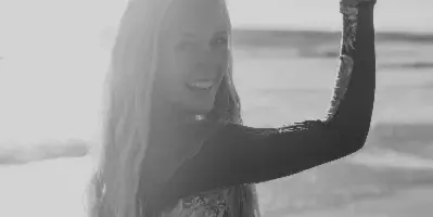 Black and white portrait of a smiling woman with long hair standing on a beach, holding onto a wooden railing.