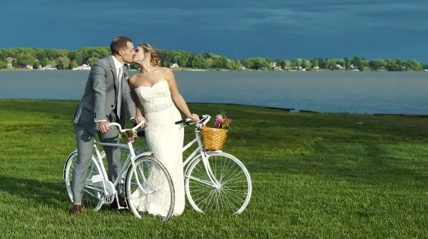 A bride and groom kissing next to white bicycles on a grassy field by a lake.