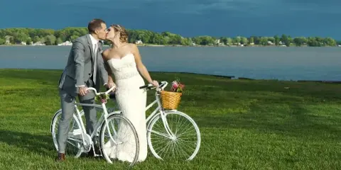 A bride and groom kissing next to white bicycles on a grassy field by a lake.