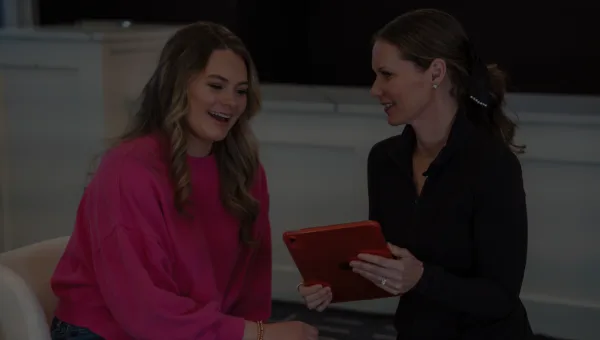 A medical professional in a black top showing a tablet to a smiling client in a pink sweatshirt during a consultation.