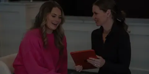 A medical professional in a black top showing a tablet to a smiling client in a pink sweatshirt during a consultation.