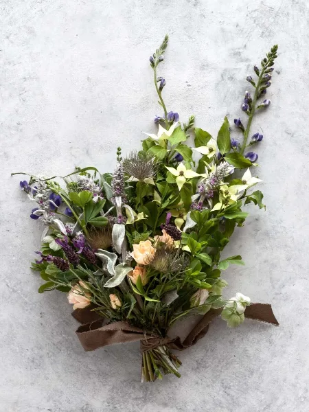 A hand-tied floral bouquet with purple and white flowers, greenery, and a brown ribbon, set against a light grey background.