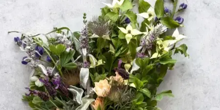 A hand-tied floral bouquet with purple and white flowers, greenery, and a brown ribbon, set against a light grey background.