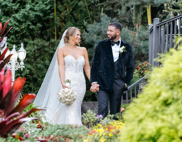 A bride in a white lace gown and a groom in a black tuxedo walking hand-in-hand through a lush garden.