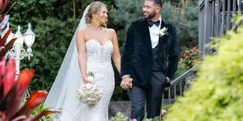 A bride in a white lace gown and a groom in a black tuxedo walking hand-in-hand through a lush garden.
