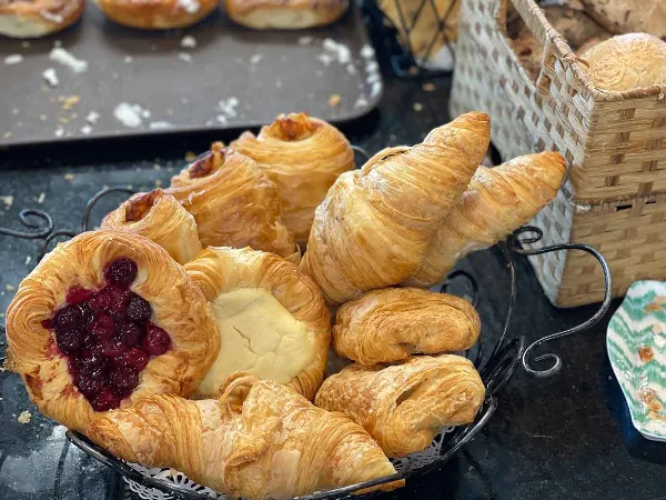A wire basket filled with assorted baked pastries, including croissants and fruit-filled danishes, displayed in a bakery setting.