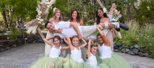 A wedding party including a groom, bridesmaids, and flower girls posing outdoors in a garden setting.