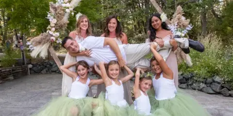 A wedding party including a groom, bridesmaids, and flower girls posing outdoors in a garden setting.