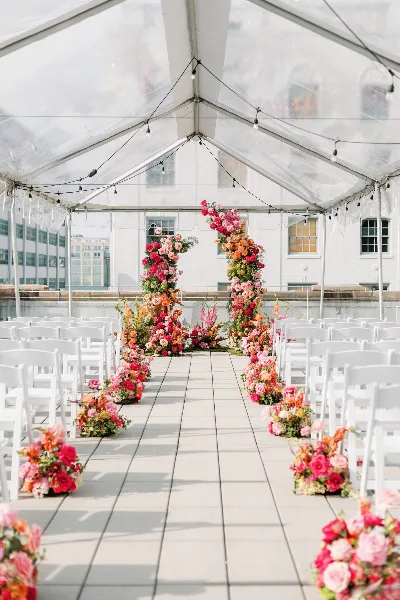 A wedding ceremony setup under a clear tent featuring a vibrant floral arch and aisle arrangements.