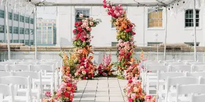 A wedding ceremony setup under a clear tent featuring a vibrant floral arch and aisle arrangements.