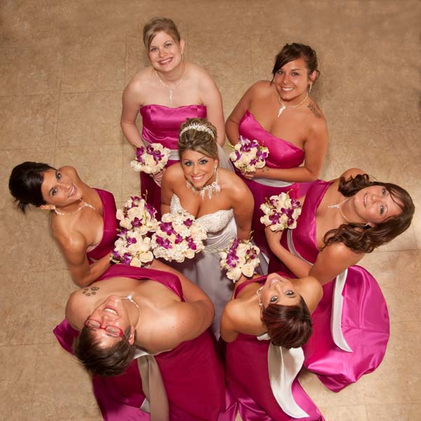 A bride in a white gown surrounded by bridesmaids in pink dresses, holding bouquets, photographed from above.