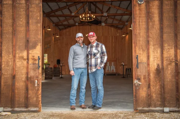 Two men standing in the open doorway of a rustic wooden barn venue with a modern chandelier and concrete floor.