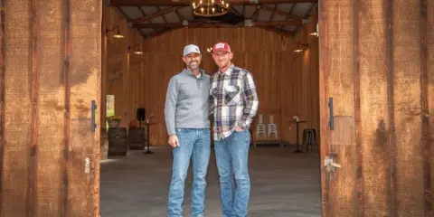 Two men standing in the open doorway of a rustic wooden barn venue with a modern chandelier and concrete floor.