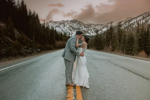 A bride and groom kissing on a paved road with snow-capped mountains and pine trees in the background.