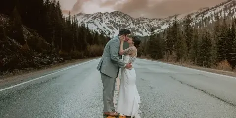 A bride and groom kissing on a paved road with snow-capped mountains and pine trees in the background.