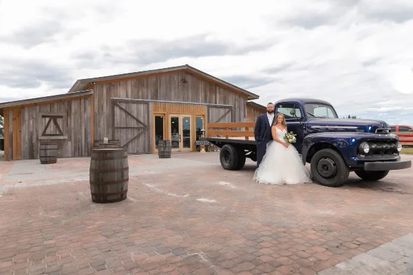 A bride and groom standing in front of a rustic wooden barn venue with a vintage blue truck and wooden barrels.