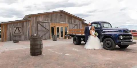 A bride and groom standing in front of a rustic wooden barn venue with a vintage blue truck and wooden barrels.