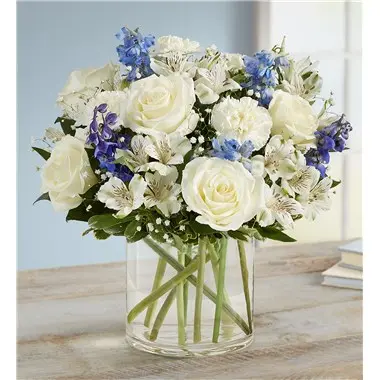 A floral arrangement of white roses and blue alstroemeria in a clear glass vase on a wooden table.