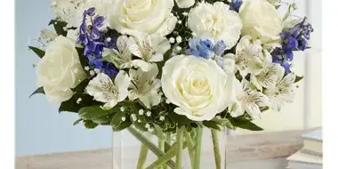 A floral arrangement of white roses and blue alstroemeria in a clear glass vase on a wooden table.
