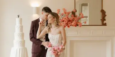 A bride and groom standing together in a bright, elegant room with a wedding cake and floral arrangements.