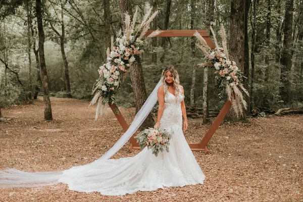 A bride in a white lace gown and long veil stands in a forest clearing in front of a wooden arch decorated with white and pink flowers and pampas grass.