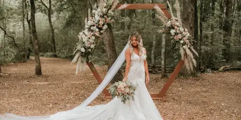 A bride in a white lace gown and long veil stands in a forest clearing in front of a wooden arch decorated with white and pink flowers and pampas grass.