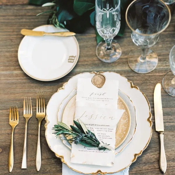 A wedding table setting featuring vintage gold-rimmed china plates, crystal wine glasses, gold flatware, and a floral centerpiece on a rustic wooden table.