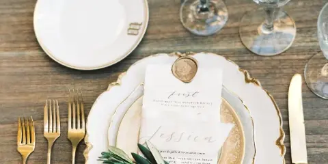 A wedding table setting featuring vintage gold-rimmed china plates, crystal wine glasses, gold flatware, and a floral centerpiece on a rustic wooden table.