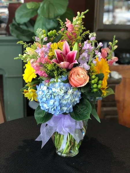 A colorful floral arrangement in a glass vase featuring pink lilies, peach roses, blue hydrangeas, and yellow flowers, tied with a purple ribbon.