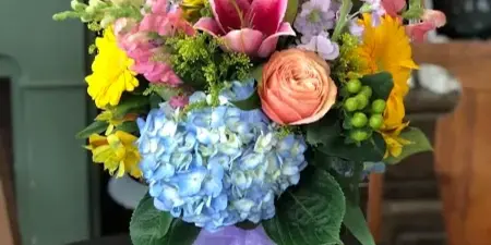 A colorful floral arrangement in a glass vase featuring pink lilies, peach roses, blue hydrangeas, and yellow flowers, tied with a purple ribbon.