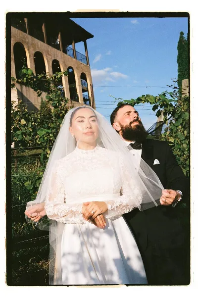A film-style portrait of a bride and groom standing together outdoors near a building with arched windows.