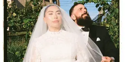 A film-style portrait of a bride and groom standing together outdoors near a building with arched windows.