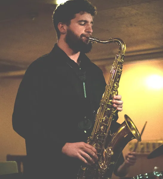 A bearded musician playing a saxophone on stage with a drummer visible in the background.