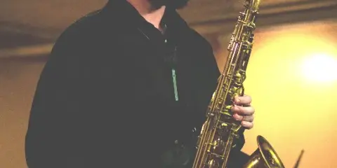 A bearded musician playing a saxophone on stage with a drummer visible in the background.